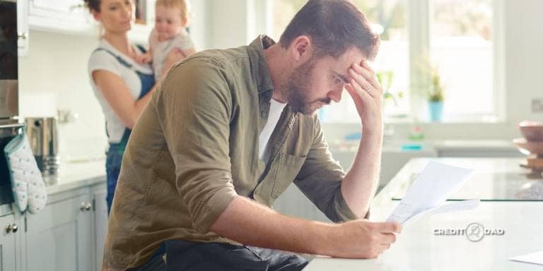 A father worried about holiday debt payoff while reviewing bills at the kitchen table with his family in the background.